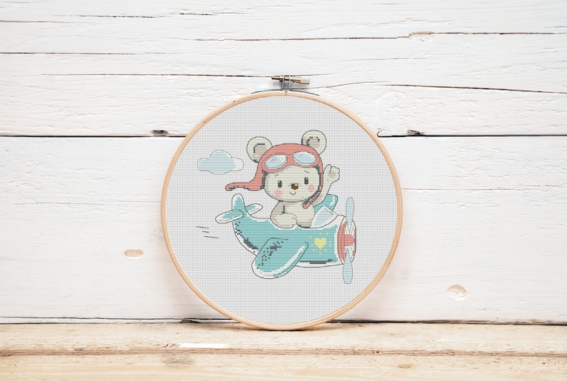 May include: A white teddy bear wearing a red aviator hat and goggles is flying a blue aeroplane with yellow accents. The aeroplane has a heart on the side. The image is a cross-stitch pattern.