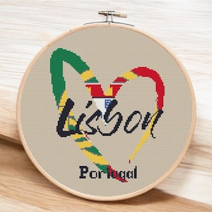 May include: A cross-stitch embroidery hoop with a heart-shaped design featuring the Portuguese flag colors and the word "Lisbon" in black cursive lettering. The text "Portugal" is also included below the heart.