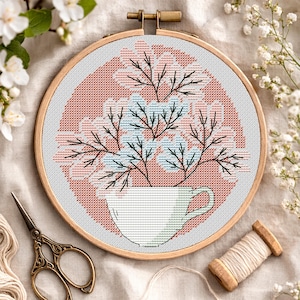 May include: Cross-stitch embroidery in a wooden hoop. The design features a white teacup filled with pink and blue flowers and black branches, set against a peach-colored background. A pair of gold scissors and a spool of thread are visible.