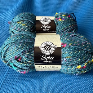 Puede incluir: Dos madejas de hilo color turquesa con motas rosas, amarillas y de otros colores. El hilo está etiquetado como "Spice Fashion" por Loops & Threads. Cada madeja contiene 140 metros de hilo.