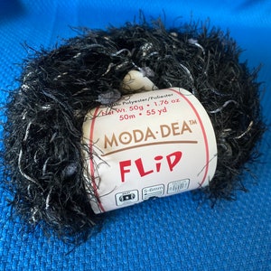 Pode incluir: Uma bola de fio preto e branco Moda Dea Flip. O rótulo diz "Flip" e inclui o peso, a composição da fibra e o tamanho da agulha recomendado. O fio tem 50g, 1,76 oz e 50 metros.