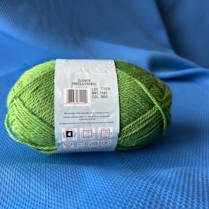 Puede incluir: Una madeja de hilo Clover de color verde brillante con una etiqueta que dice "CLOVER TRÈFLE/TRÉBOL". La etiqueta incluye detalles sobre el hilo. El hilo es de grosor medio y adecuado para varios proyectos de tejido y ganchillo. Mide 10 x 10 cm.