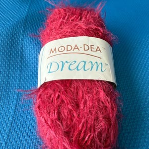 Pode incluir: Um novelo de fio fúcsia vibrante com uma textura fofa. O fio é rotulado "MODA-DEA Dream" em um design branco e azul. O fio está sobre um fundo azul texturizado.