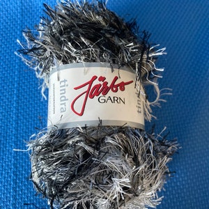 Può includere: Un gomitolo di filato nero e bianco soffice con un'etichetta che recita "Järbo Garn" e "Tindra" in testo bianco su sfondo rosso.