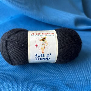 Puede incluir: Una madeja de lana negra con una etiqueta que dice "Stitch Nation by Debbie Stoller" y "full o' sheep". La etiqueta presenta una ilustración retro de una mujer en traje de baño. La lana está sobre un fondo de tela azul.