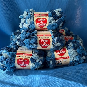 May include: A stack of blue and white Pomp-a-Doodle yarn balls. Each ball has a Red Heart label. The yarn is a bulky weight and has a textured, pom-pom-like appearance. The background is a solid blue.