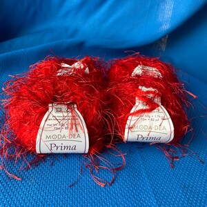 Pode incluir: Dois novelos de fio vermelho felpudo com etiquetas brancas que dizem "Moda Dea Prima" e incluem o peso, o comprimento e o conteúdo da fibra.