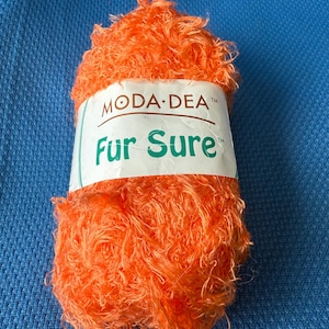 Pode incluir: Novel de lã laranja brilhante com aparência felpuda. A etiqueta, em branco e verde, diz "MODA-DEA Fur Sure". Adequado para projetos de tricô ou crochê.