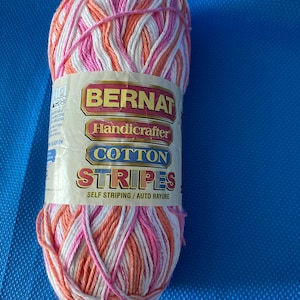 Op de afbeelding: Een bol Bernat Handicrafter Cotton Stripes garen in een zelfstreepend patroon van roze, wit en oranje.