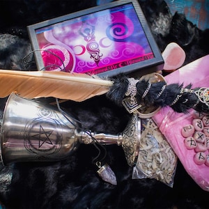 May include: A silver chalice with a pentagram design, a feather, a black and silver beaded wand, a small bag of white sage, a pink bag with runes, and a pink box with the text "Blessed Be" on the lid.