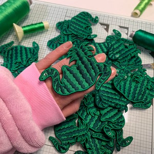 May include: A collection of embroidered patches featuring a green and dark green striped serpentine creature design. The patches are displayed alongside spools of green thread, gold-coloured scissors, and a white cutting mat with grid lines.