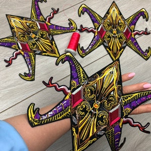 May include: Three star-shaped embroidered patches featuring intricate gold, purple, and black patterns with red diamond-shaped accents. The designs include swirling filigree details and curved points. A small spool of red thread sits between the patches.