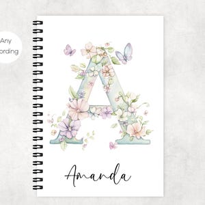 Può includere: Un quaderno a spirale con un design floreale. La copertina presenta una grande lettera "A" adornata con fiori e farfalle dai colori pastello. Il nome "Amanda" è scritto in corsivo sotto.