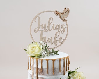 May include: A white cake with gold drip icing and white flowers. A wooden sign with the text "Julias Taufe" and a dove with an olive branch is on top of the cake.