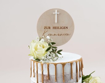 Op de afbeelding: Een taart versierd met witte glazuur en gouden druppels. Een houten schijf met een wit kruis en de tekst "ZUR HEILIGEN KOMMUNION" staat erop. De taart is versierd met witte rozen en groen.