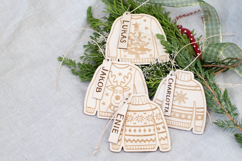 Peut inclure: Quatre d&eacute;corations de No&euml;l en bois en forme de pulls avec des motifs diff&eacute;rents. Chaque d&eacute;coration a une &eacute;tiquette en bois avec un nom dessus : "Lukas", "Jakob", "Charlotte" et "Elie".