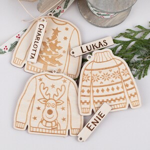 Peut inclure: Trois d&eacute;corations de No&euml;l en bois en forme de pulls avec des motifs diff&eacute;rents. Chaque d&eacute;coration a une &eacute;tiquette avec un nom : Charlotte, Lukas et Enie.