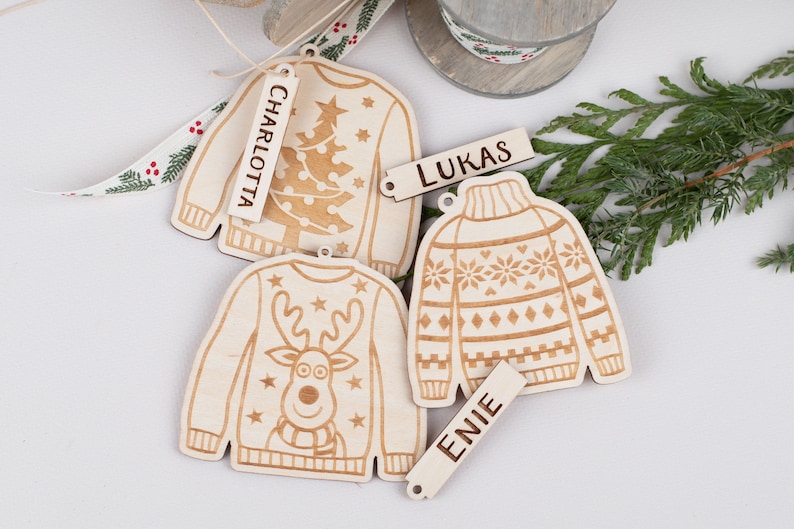 Peut inclure: Trois d&eacute;corations de No&euml;l en bois en forme de pulls avec des motifs diff&eacute;rents. Chaque d&eacute;coration a une &eacute;tiquette avec un nom : Charlotte, Lukas et Enie.
