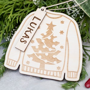 Peut inclure: Une d&eacute;coration de No&euml;l en bois en forme de pull avec un motif de sapin de No&euml;l. L'ornement a une &eacute;tiquette attach&eacute;e qui dit "Lukas".