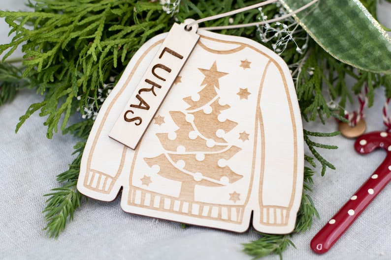 Peut inclure: Une d&eacute;coration de No&euml;l en bois en forme de pull avec un motif de sapin de No&euml;l. L'ornement a une &eacute;tiquette attach&eacute;e qui dit "Lukas".