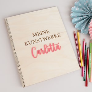 Op de afbeelding: Een houten doos met de tekst "Meine Kunstwerke Carlotta" gegraveerd op de voorkant. De doos is omringd door kleurpotloden en papieren waaier.