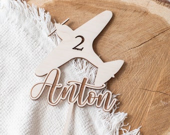 May include: Wooden airplane cake topper with the name Anton and the number 2. The topper is on a white fabric background.