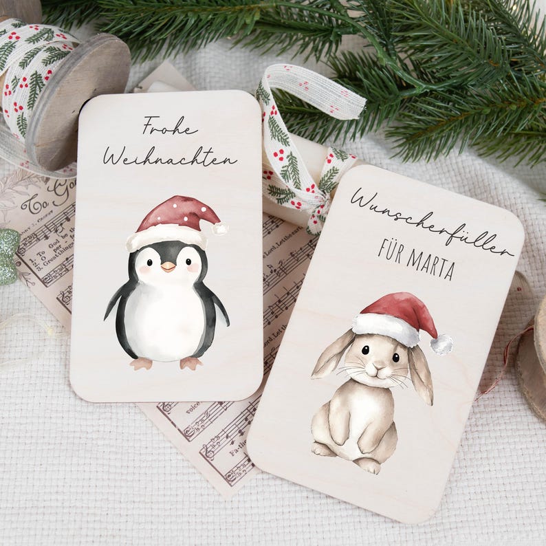 May include: Two wooden Christmas gift tags with watercolor illustrations. One tag features a penguin wearing a Santa hat with the text "Frohe Weihnachten". The other tag features a bunny wearing a Santa hat with the text "Wunschersch&uuml;ller f&uuml;r Marta".