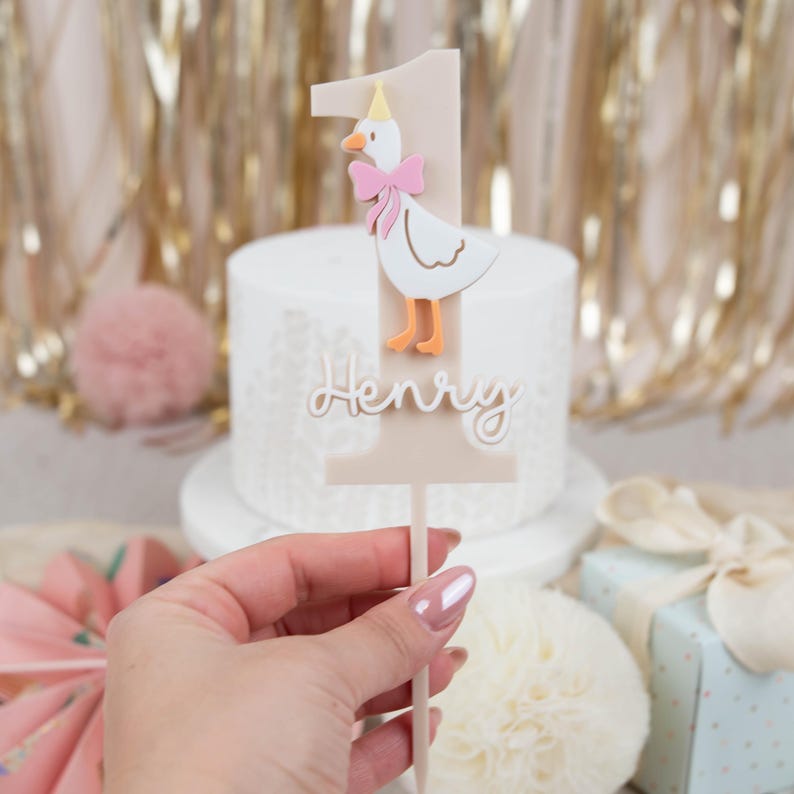 May include: A beige cake topper featuring a white goose wearing a pink bow and a yellow party hat. The topper is shaped like the number one and has the name "Henry" written on it.