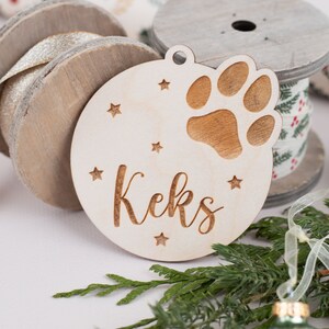 May include: A wooden Christmas ornament with a paw print and the name "Keks" engraved on it. The ornament is surrounded by green pine branches and a silver ribbon.