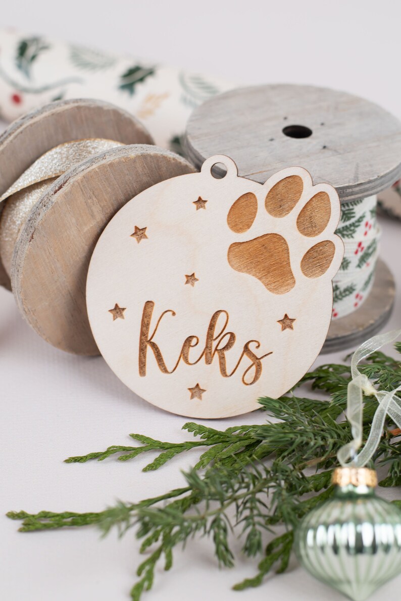 May include: A wooden Christmas ornament with a paw print and the name "Keks" engraved on it. The ornament is surrounded by green pine branches and a silver ribbon.