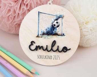 Pode incluir: Enfeite redondo de madeira com uma bola de futebol numa rede e detalhes em aquarela azul. O nome "Emilio" está escrito em itálico preto, com "SCHULKIND 2025" por baixo. Possui um cordão para pendurar.