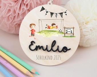Pode incluir: Enfeite redondo de madeira com tema de futebol. Apresenta uma baliza de futebol, um jogador e o nome "Emilio" em escrita preta. O texto "SCHULKIND 2025" também está presente.