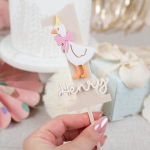 May include: A beige cake topper shaped like the number one with a white duck wearing a pink bow and a yellow party hat. The name "Henry" is written in white on the topper.