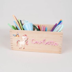 Peut inclure: Une boîte à crayons en bois remplie de stylos, crayons et un taille-crayon bleu. La boîte est décorée d'une licorne et d'un papillon, avec le nom "Carlotte" en rose.