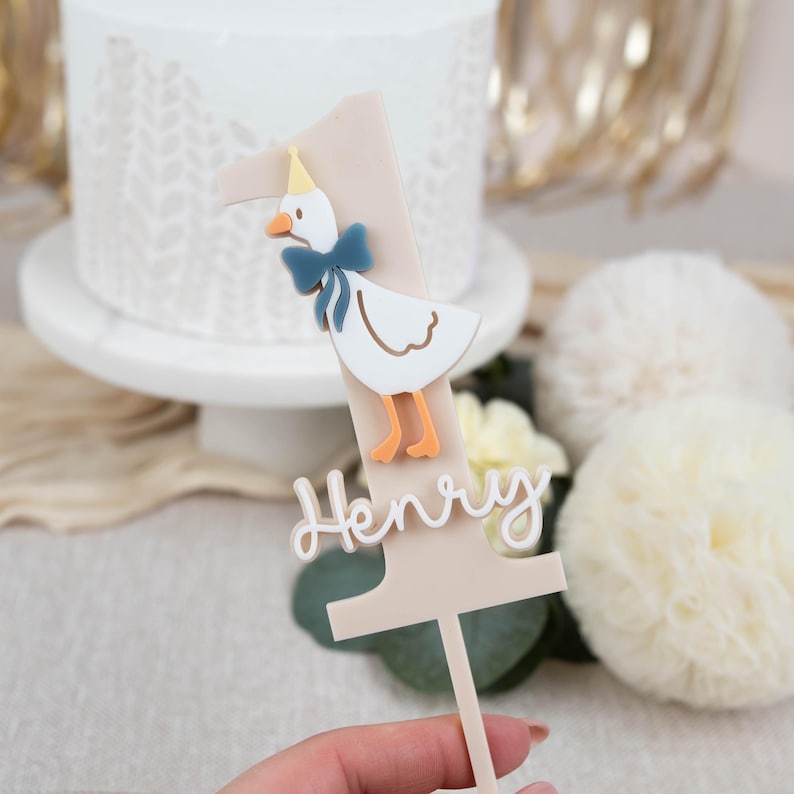 May include: A beige acrylic cake topper shaped like the number one with a white duck wearing a yellow party hat and a blue bow tie. The name "Henry" is written in cursive on the topper.