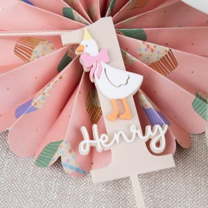 May include: A white cake topper featuring a pink and white duck wearing a party hat. The topper is decorated with the number one and the name "Henry" in white.