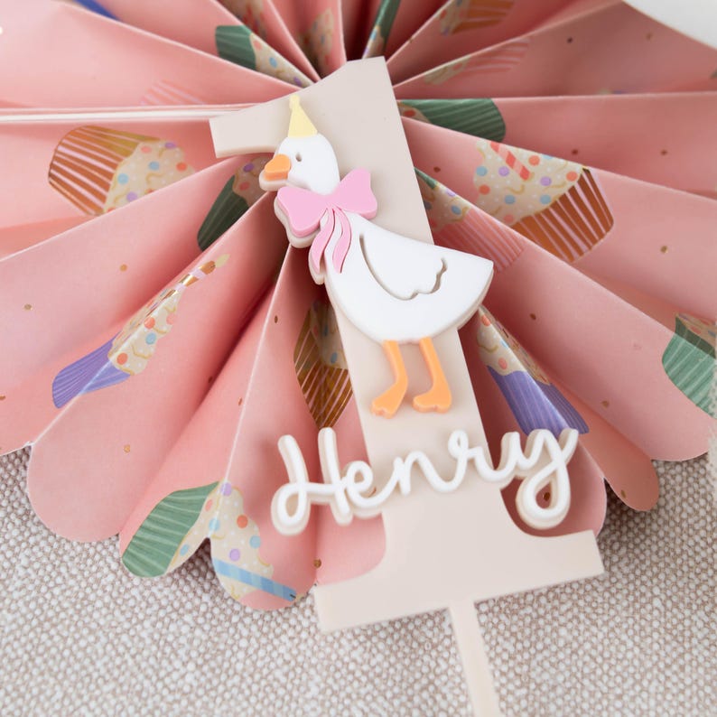 May include: A white cake topper featuring a pink and white duck wearing a party hat. The topper is decorated with the number one and the name "Henry" in white.