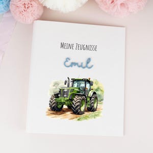 Könnte beinhalten: Ein weißer, rechteckiger Ordner mit dem schwarzen Schriftzug "Meine Zeugnisse", dem Namen "Emil" in blauer Schreibschrift und einer Aquarellillustration eines grünen Traktors auf einem Feldweg mit grünen Bäumen.