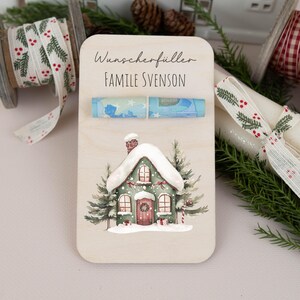 May include: A wooden Christmas money holder with a watercolor illustration of a snow-covered house. The holder has a slot for cash and the text "Wunscherschiller Familie Svenson".