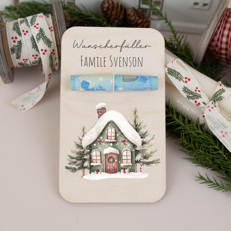 May include: A wooden Christmas money holder with a watercolor illustration of a snow-covered house. The holder has a slot for cash and the text "Wunscherschiller Familie Svenson".