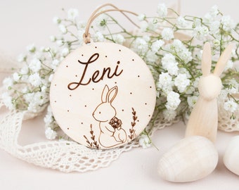 Op de afbeelding: Een ronde houten hanger met de naam "Leni" erop gegraveerd. Een klein konijntje met een bloemetje is ook gegraveerd op de hanger. De hanger hangt aan een touwtje en is omringd door witte bloemen.