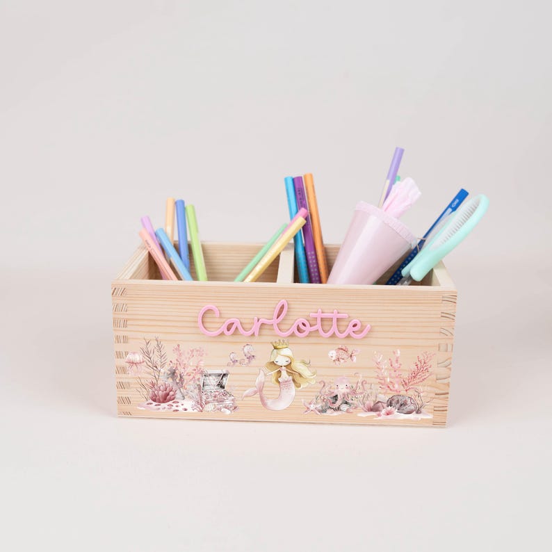 May include: A wooden desk organizer with a mermaid theme. The organizer is filled with pens, pencils, and a pink cup. The name "Charlotte" is displayed in pink letters on the front, with a mermaid and underwater scene.