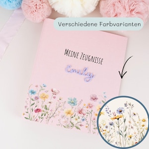 Peut inclure: Un dossier rectangulaire rose clair portant le texte MEINE ZEUGNISSE et le prénom Emily en écriture cursive violette. La partie inférieure est ornée d'une illustration aquarelle colorée de fleurs sauvages dans des tons bleus, jaunes et roses.
