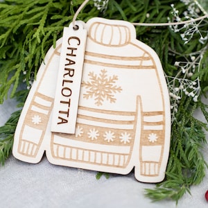 Peut inclure: Une d&eacute;coration de No&euml;l en bois en forme de pull avec un motif flocon de neige. L'ornement a une &eacute;tiquette attach&eacute;e qui dit "CHARLOTTA".