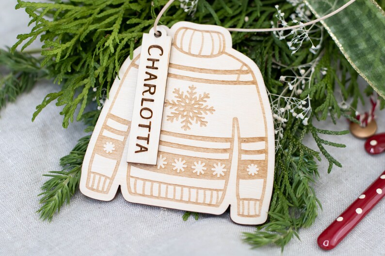 Peut inclure: Une d&eacute;coration de No&euml;l en bois en forme de pull avec un motif flocon de neige. L'ornement a une &eacute;tiquette attach&eacute;e qui dit "CHARLOTTA".