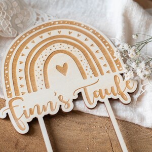 May include: Wooden rainbow cake topper with the text "Finn's Taufe". The topper features a rainbow design with heart and dot details. The cake topper is made of light-colored wood and has two sticks for insertion.