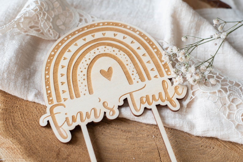 May include: Wooden rainbow cake topper with the text "Finn's Taufe". The topper features a rainbow design with heart and dot details. The cake topper is made of light-colored wood and has two sticks for insertion.
