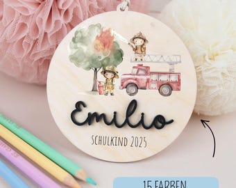 Pode incluir: Enfeite redondo de madeira com uma ilustração em aquarela de bombeiros e um carro de bombeiros. O nome "Emilio" está escrito em itálico preto, com "SCHULKIND 2025" abaixo. O enfeite tem uma corda para pendurar e o texto "15 FARBEN" é visível.