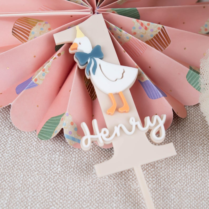 May include: A white cake topper with a pink number one, a white goose with a blue bow, and the name "Henry" in white script.