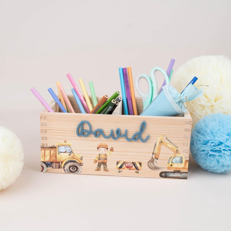 May include: A rectangular wooden desk organiser featuring the name David in blue script. The front displays watercolour illustrations of a yellow dump truck, a construction worker, a road sign, and a yellow excavator, filled with various coloured pencils.